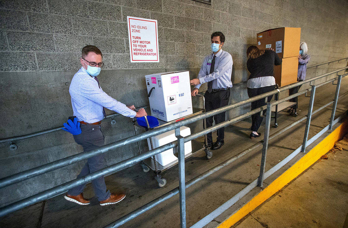 University of Washington Medical Center Montlake campus pharmacy administration resident Derek Pohlmeyer (left) and UWMC pharmacy director Michael Alwan transport a box containing Pfizer-BioNTech COVID-19 vaccines toward a waiting vehicle headed to the UW Medical Centers other hospital campuses on Monday in Seattle. (Mike Siegel/The Seattle Times via media pool)